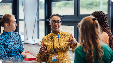 A group of women engaged in conversation in a classroom setting, surrounded by desks and educational materials.